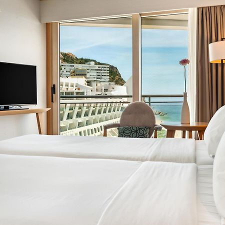 Sesimbra Hotel&Spa Exterior foto The photo shows a hotel room featuring two neatly made beds with white linens. There is a small table with a chair, and a television mounted on a wooden stand. The large window provides a view of a coastal scene, with buildings on a hillside and wate