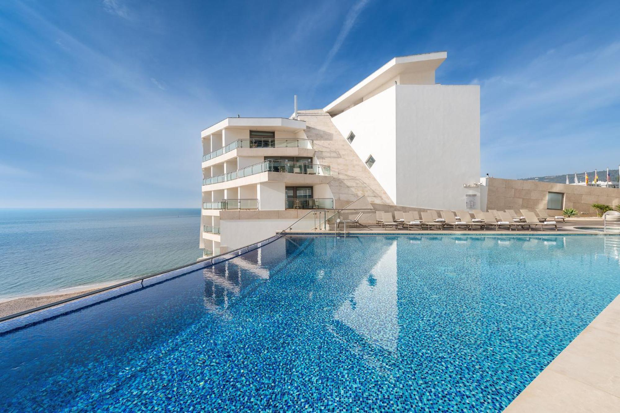 Sesimbra Hotel&Spa Exterior foto The photo shows a modern hotel building located by the seaside. In the foreground, there is an expansive infinity pool with a clear blue surface that reflects the sky. The pool appears to extend towards the ocean, creating a seamless view. The hotel 