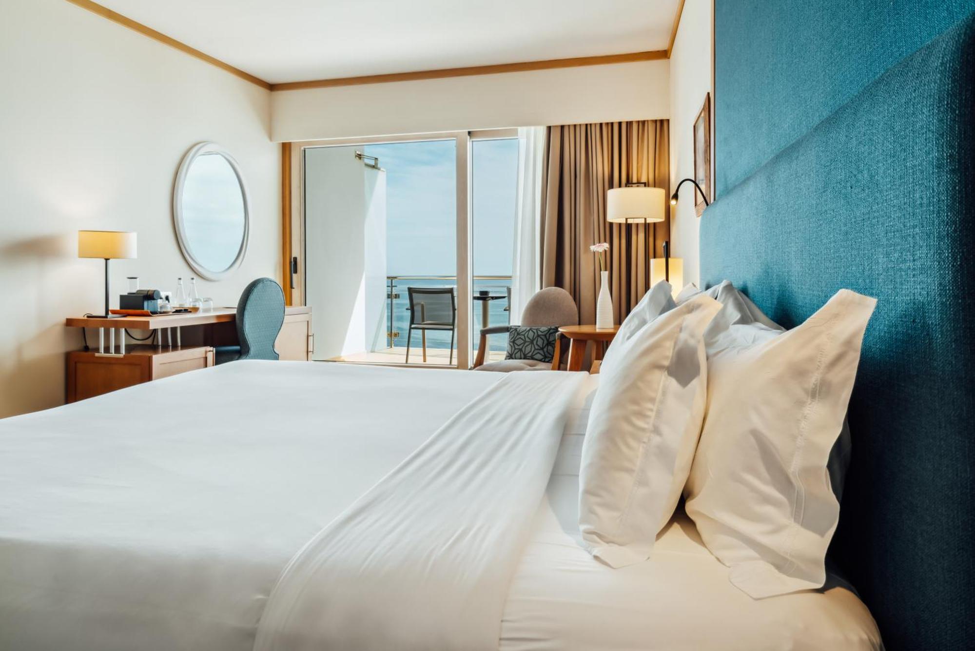 Sesimbra Hotel&Spa Exterior foto The photo shows a well-appointed hotel room featuring a large bed with plush white linens and pillows. The wall behind the bed is a soft blue fabric, creating a calming atmosphere. There is a round mirror on the wall and a stylish desk area to the le