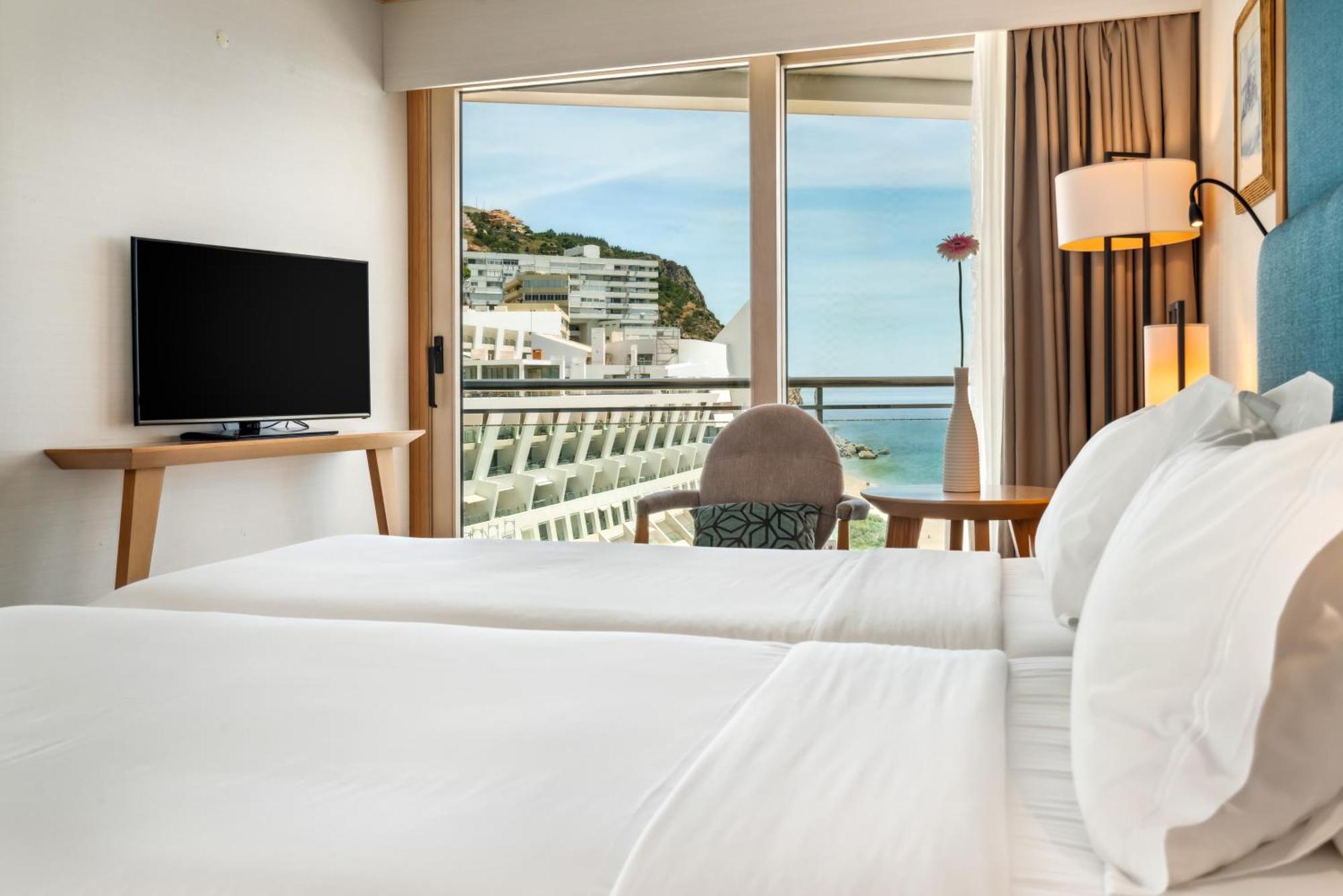 Sesimbra Hotel&Spa Exterior foto The photo shows a hotel room featuring two neatly made beds with white linens. There is a small table with a chair, and a television mounted on a wooden stand. The large window provides a view of a coastal scene, with buildings on a hillside and wate
